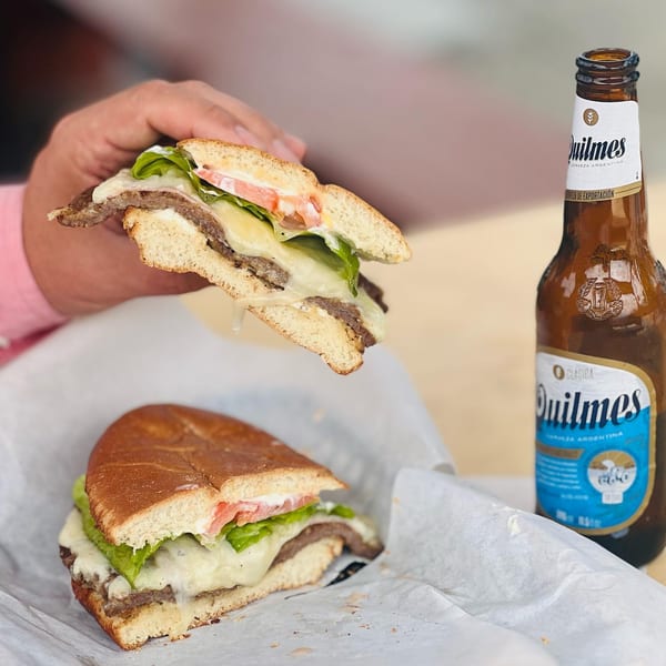 A hand holds an Argentinian lomito sandwich with melted cheese, lettuce, and tomato, next to a Quilmes beer.