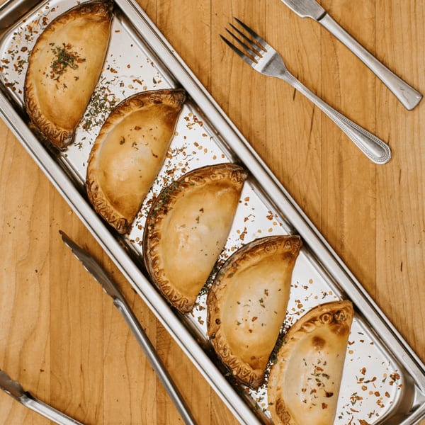 Five golden-brown JAVI'S COOKING Empanadas Argentinas on a silver tray with herbs, tongs, and cutlery.