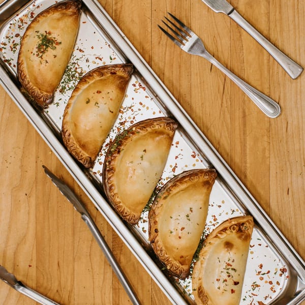 Five Javi's Cooking Empanadas Argentinas on a silver tray with spices, tongs, fork, and knife.