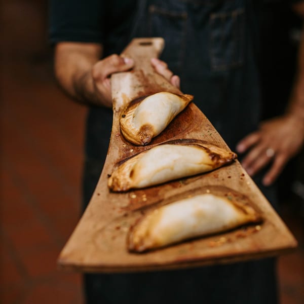 A person holds a wooden paddle with three golden-brown Argentinian empanadas, fresh from the oven.